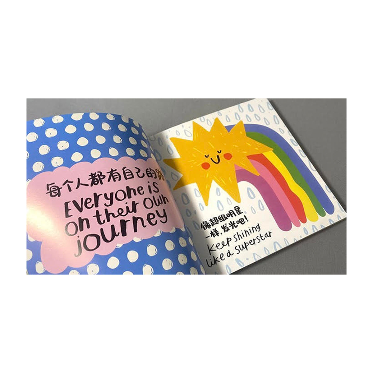 An open book with colorful pages. Left page: blue with white dots and pink cloud saying "Everyone is on their own journey" in English and Chinese. Right page: smiling sun with rainbow and "Keep shining like a superstar" in English and Chinese.