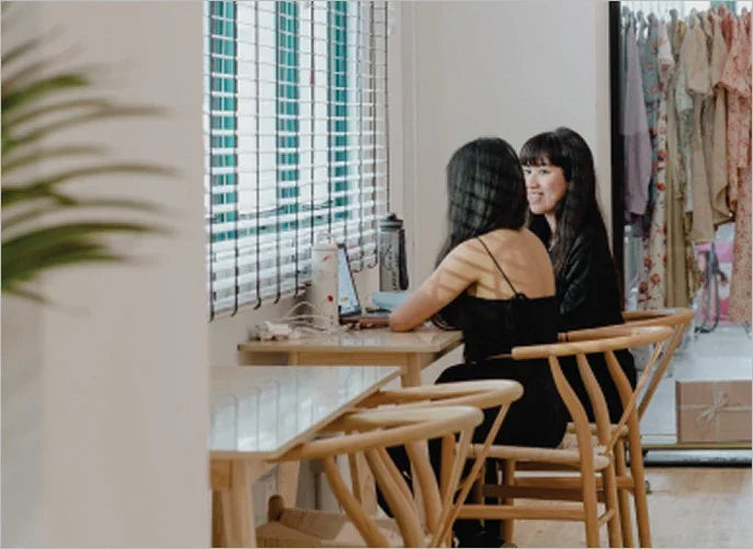 Two women sitting at a wooden table in a bright boutique near clothing racks.