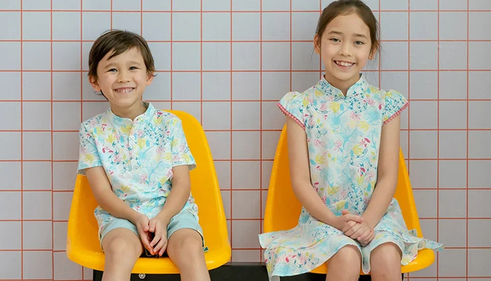 Two smiling children in matching light blue floral patterned traditional Asian outfits sitting on yellow chairs.