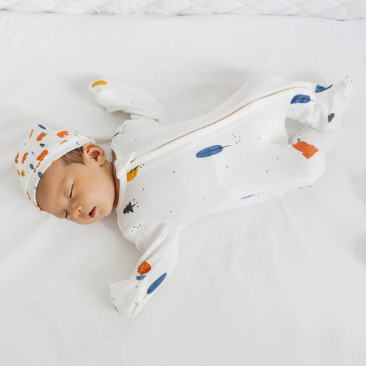 A newborn baby wearing a white, organic zip sleepsuit with foldable mittens and footies by Le Petit Society, featuring a forest motif with brown bears.