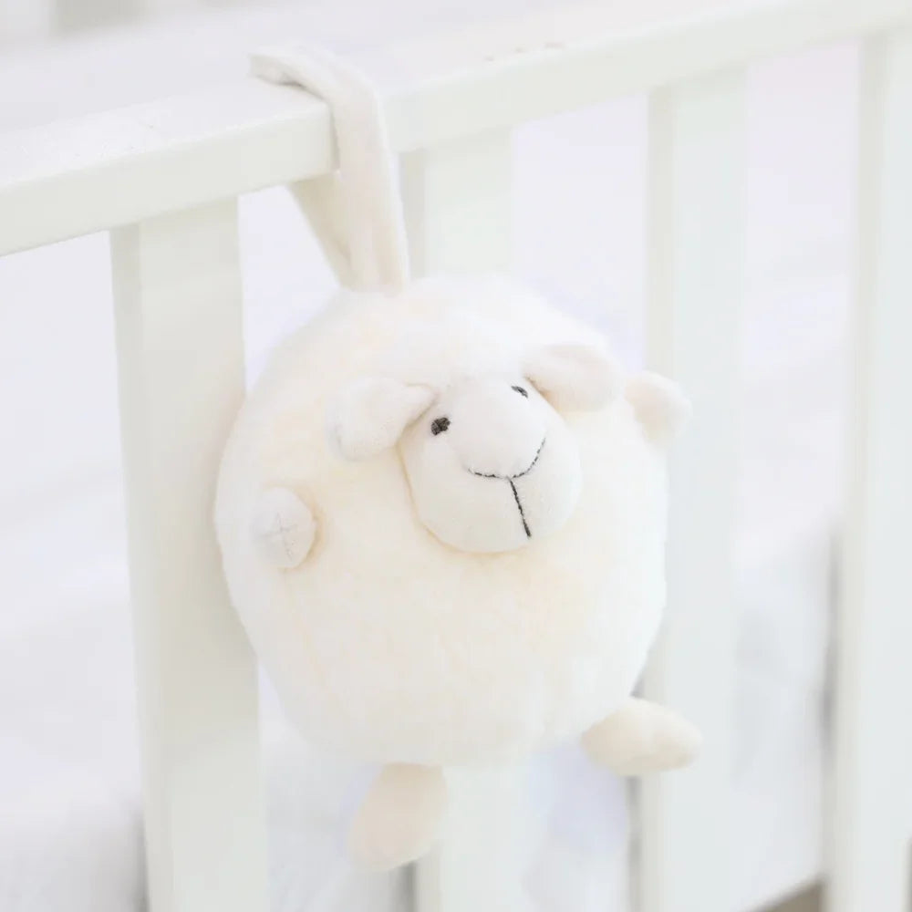 A round and plush sheep toy in cream white by Le Petit Society hangs off the frame of a baby cot.