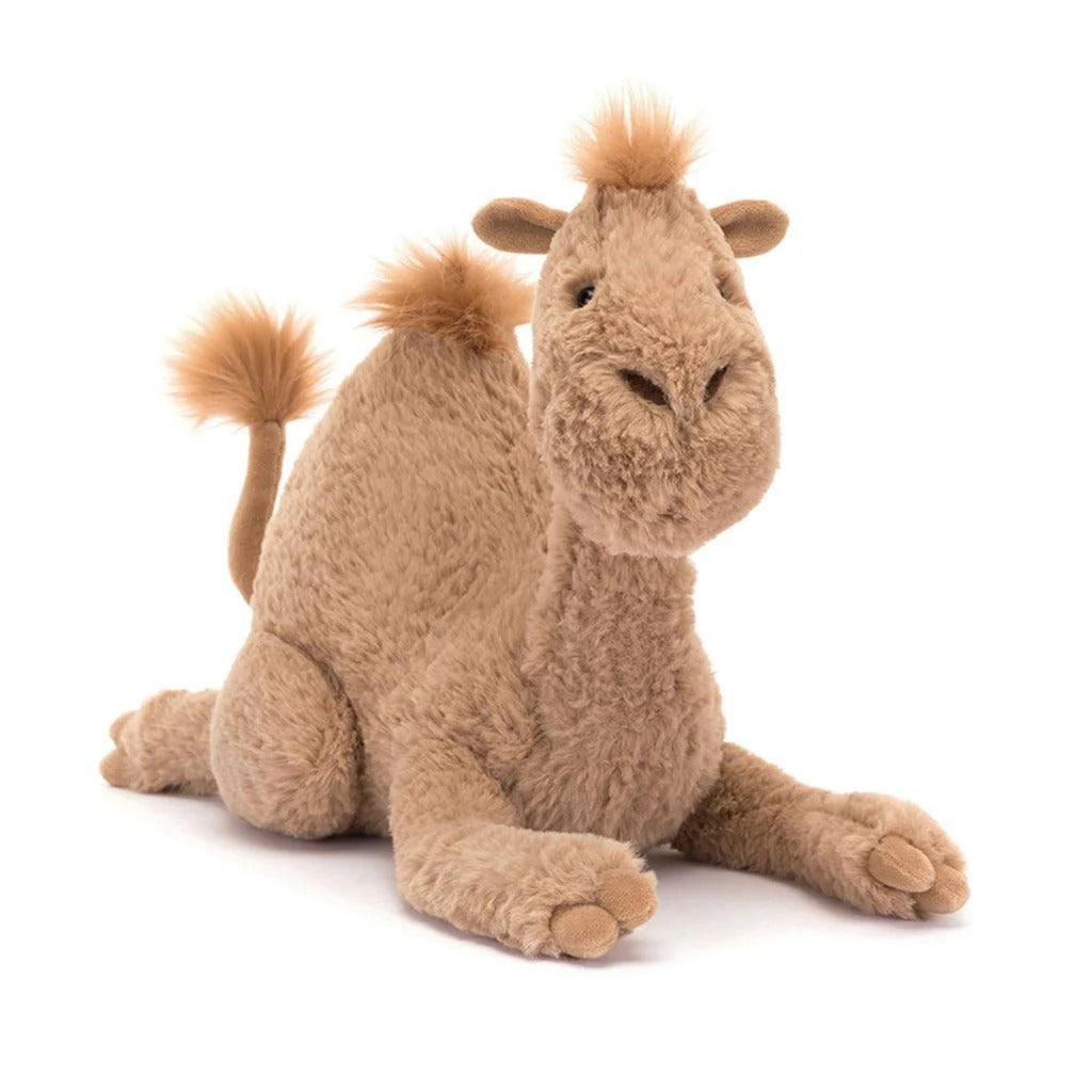A front view of a plush camel styled as Jellycat’s Richie Dromedary: soft sandy-brown fur, a pronounced hump, embroidered nostrils, a poised tail, cloven hooves, and a gentle smiling expression.