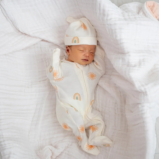 A newborn baby wearing an organic zip sleepsuit with foldable mittens and footies by Le Petit Society, featuring a print of sun and rainbows over a cream base.