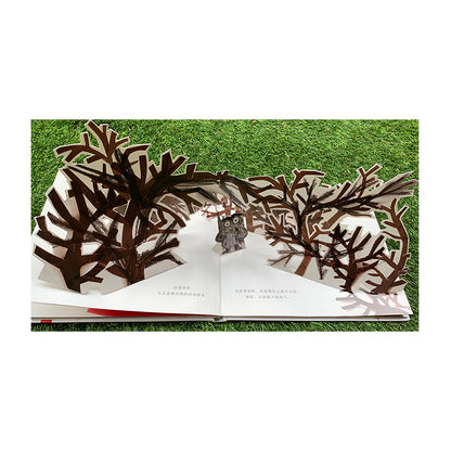 Pop-up book on grass showing a dark forest scene. Brown tree branches arch over the pages forming a tunnel. A small owl stands in the middle, surrounded by the branches. Text is visible beneath the pop-up.
