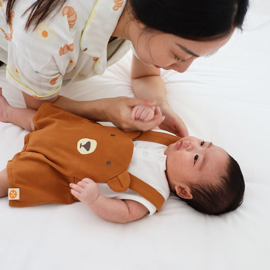 An animal-themed baby romper by Le Petit Society, featuring a brown bear's face at the front, complete with brown bottoms and brown cross back straps over a white base.