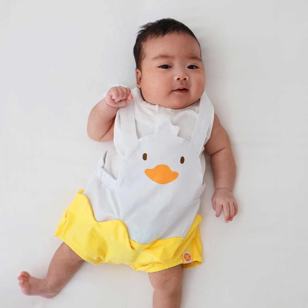 An animal-themed baby romper by Le Petit Society, featuring a duck's face at the front, complete with bright yellow bottoms and white cross back straps over a white base.