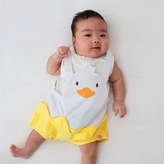 An animal-themed baby romper by Le Petit Society, featuring a duck's face at the front, complete with bright yellow bottoms and white cross back straps over a white base.