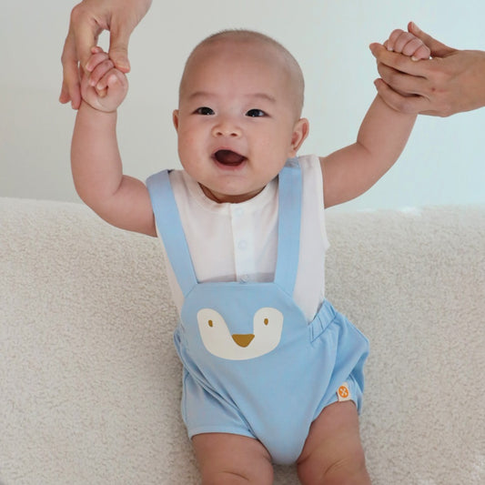 An animal-themed baby romper by Le Petit Society, featuring a blue penguin's face at the front, complete with blue bottoms and blue cross back straps over a white base.