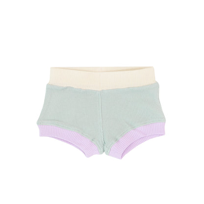 A pair of baby boxers made in a waffle jersey fabric. It has a pastel colour block colour scheme;. consisting of sage green, purple and yellow.