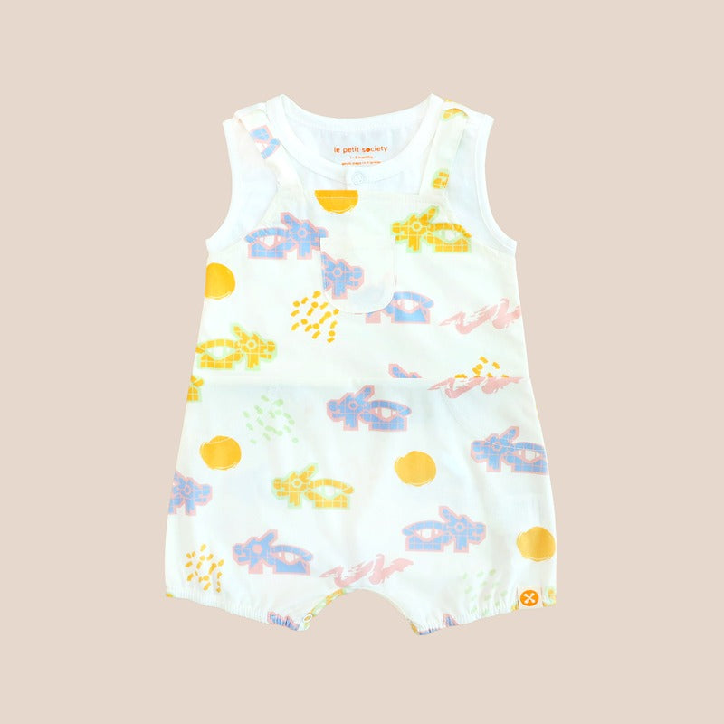 White baby overall from Le Petit Society with colorful dragon playground and sun print in pastel blue, yellow, green, and pink, featuring sleeveless straps and a front pocket.