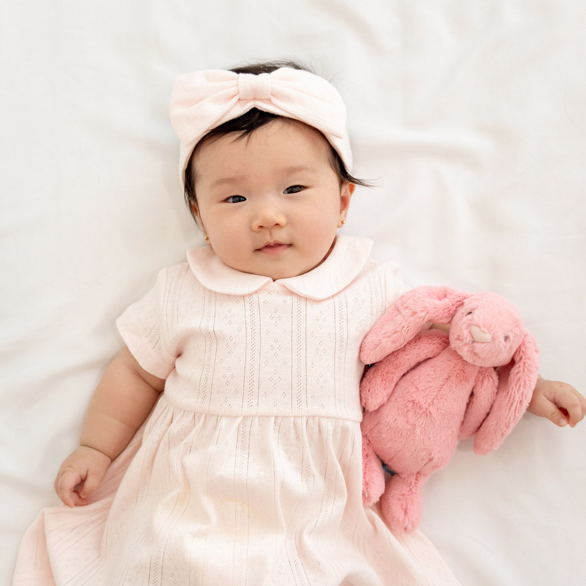 A baby girl wearing a peter pan collar dress in pink pointelle cotton with a matching bow headband from Le Petit Society.