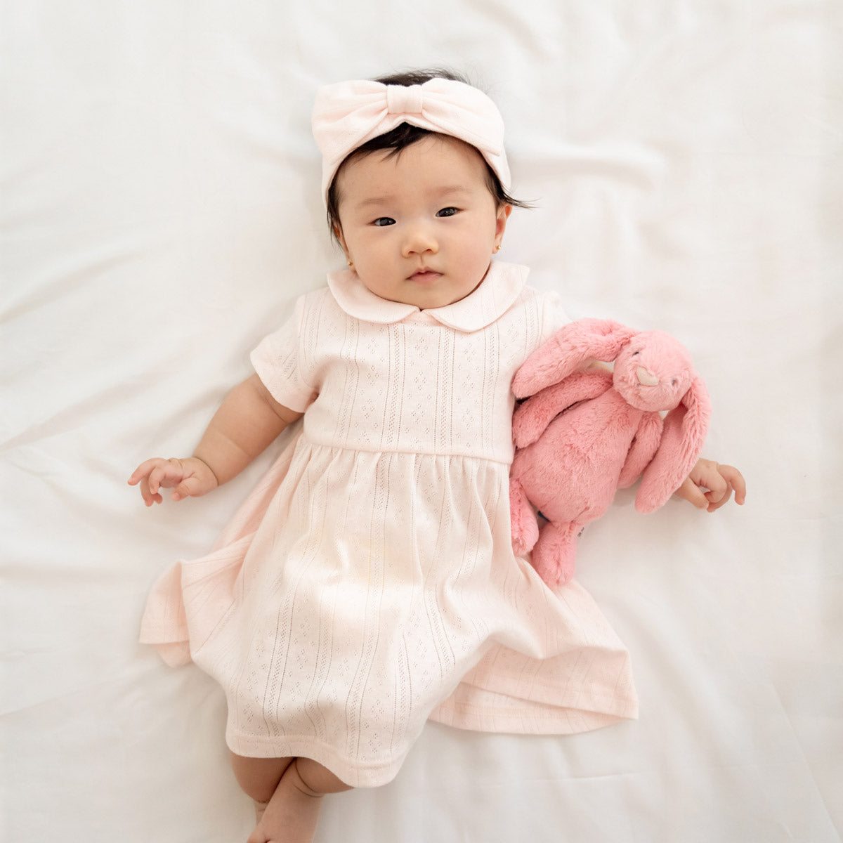 A baby girl wearing a peter pan collar dress in pink pointelle cotton with a matching bow headband.