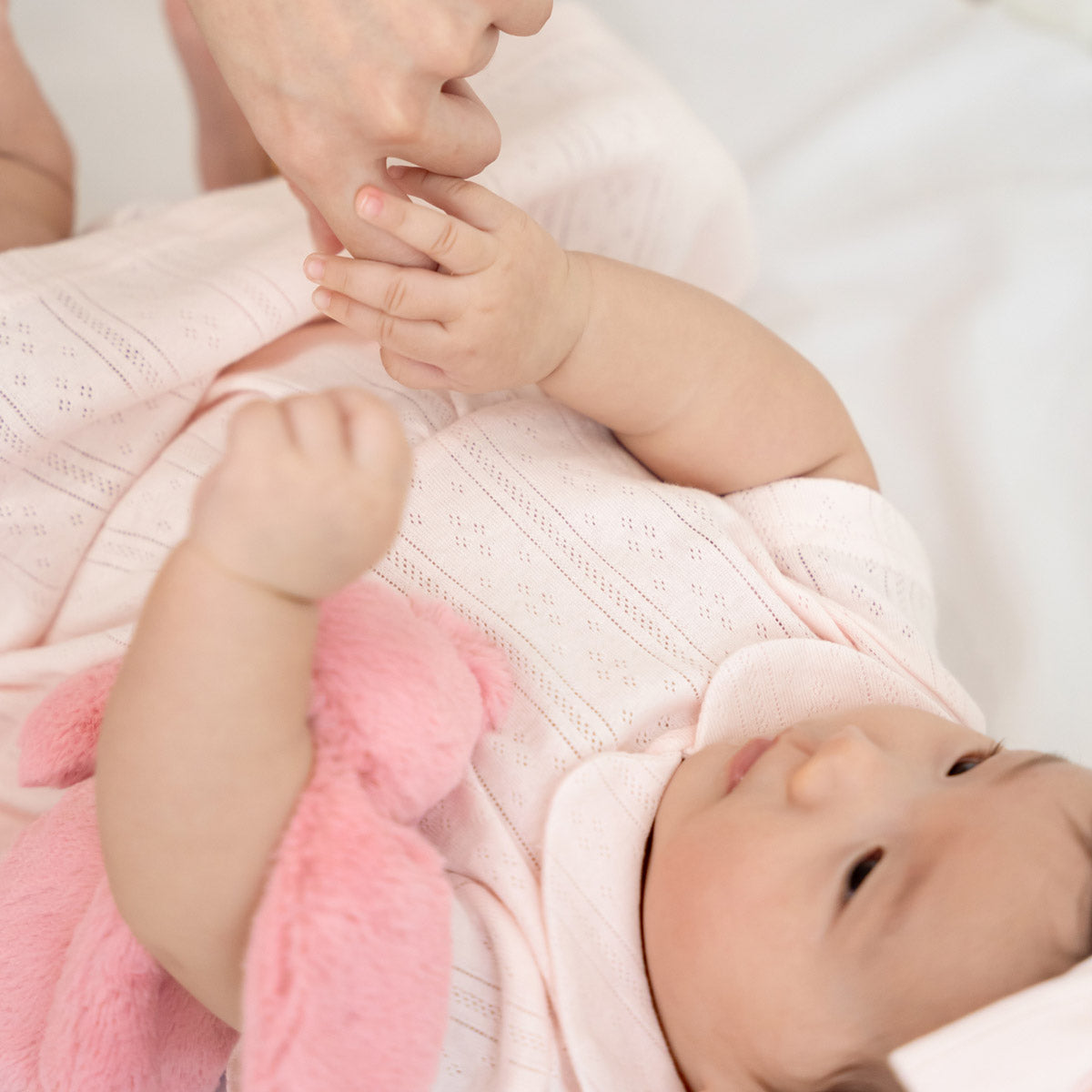 A baby girl wearing a peter pan collar dress in pink pointelle cotton with a matching bow headband.