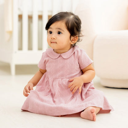 A baby girl wearing a plum-coloured, pointelle cotton dress with a peter pan collar from Le Petit Society.
