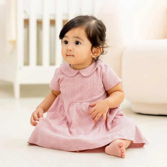 A baby girl wearing a plum-coloured, pointelle cotton dress with a peter pan collar from Le Petit Society.