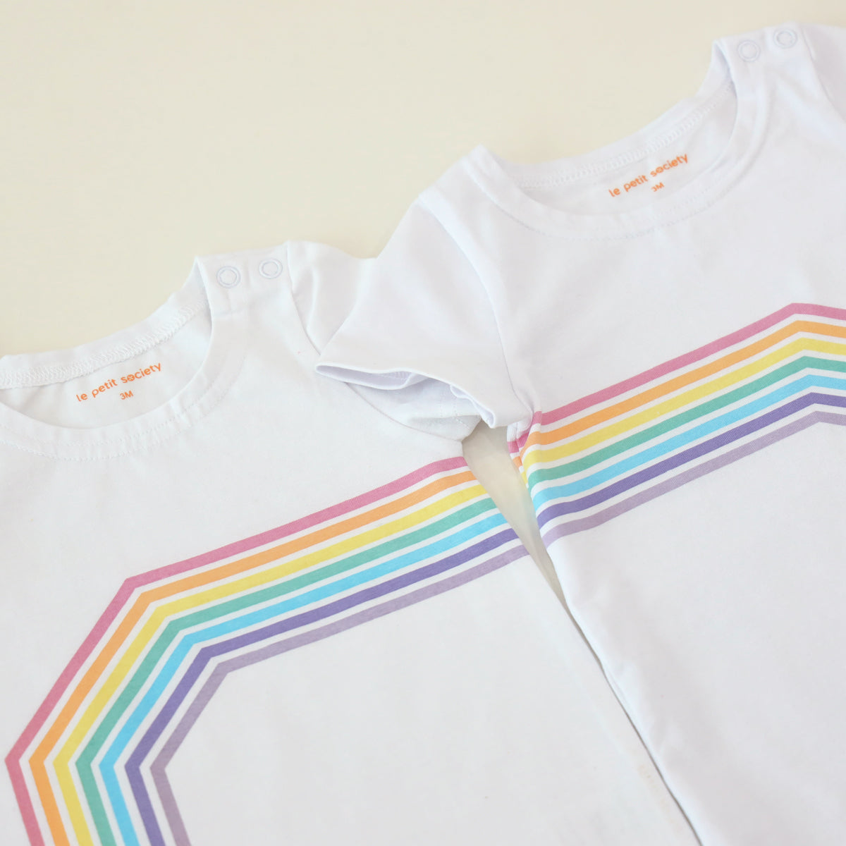Two white rompers with a print of the left and right arc of a rainbow are placed side-by-side, creating a complete rainbow.