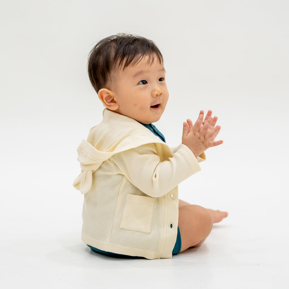 A baby boy wearing a bear cardigan in cream waffle jersey, which comes with buttons and pockets at the front, as well as a hood with little bear ears.