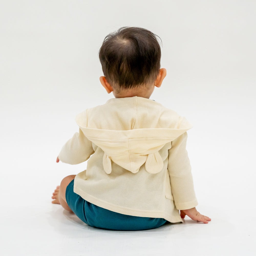 A baby boy wearing a bear cardigan in cream waffle jersey, which features a hood with little bear ears.