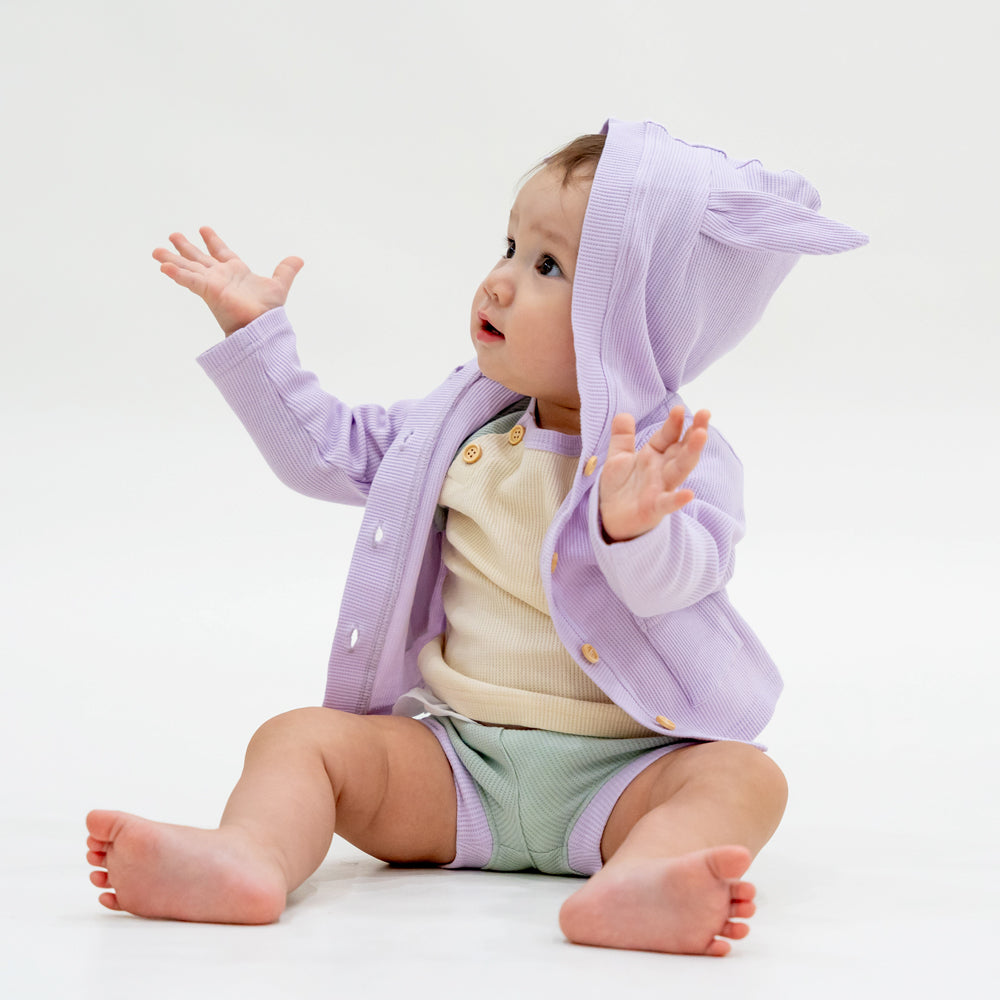 A baby boy wearing a coordinating set of a colour block tee and boxers, complete with a lilac purple bunny cardigan from Le Petit Society.