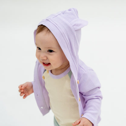 A baby boy wearing a coordinating set of a colour block tee and boxers, complete with a lilac purple bunny cardigan from Le Petit Society.