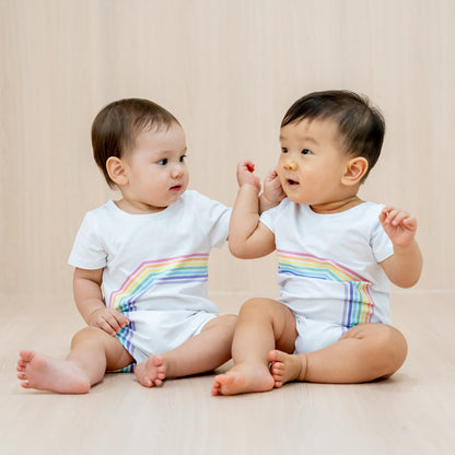 Two babies wearing white rompers with a print of the left and right arc of a rainbow respectively, a matching outfit that creates a complete rainbow when placed side-by-side.