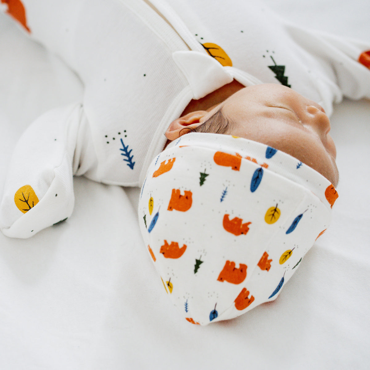 A newborn baby wearing a white, organic zip sleepsuit with foldable mittens and footies by Le Petit Society, featuring a forest motif with brown bears.