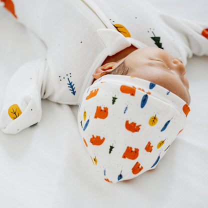 A newborn baby wearing a white, organic zip sleepsuit with foldable mittens and footies by Le Petit Society, featuring a forest motif with brown bears.