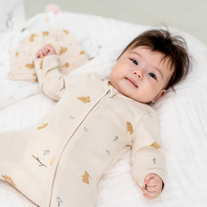 A baby wearing an organic zip sleepsuit with foldable mittens and footies by Le Petit Society, featuring a woodland-themed motif with rabbits over a beige base. 