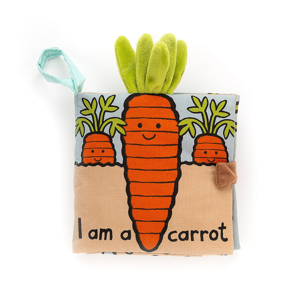 A soft fabric children's book with a smiling carrot illustration on the cover. The carrot has green plush leaves sticking out of the top. Text on the cover reads, "I am a carrot." The background is light blue and beige.