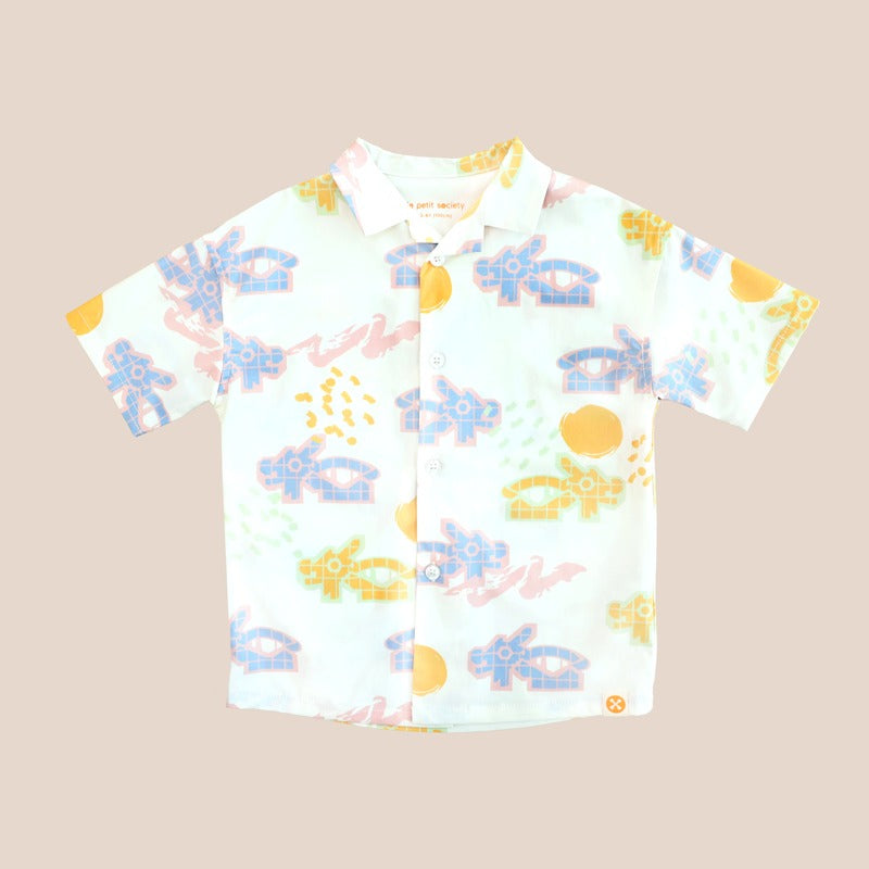 White short-sleeve boys’ button-up shirt from Le Petit Society with camp collar in colorful dragon playground print and sun print in pastel blue, yellow, green, and pink.