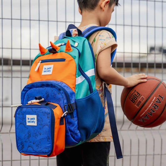 A primary school boy playing basketball while carrying the single pocket backpack in dinosaur design.
