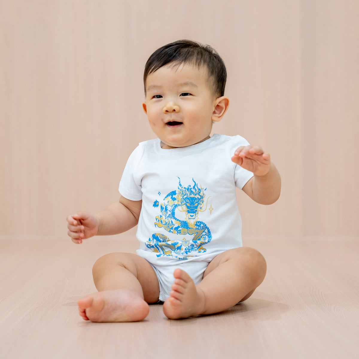 A baby boy in a white romper with a blue and gold dragon print from Le Petit Society's Dragon Streetwear collection.