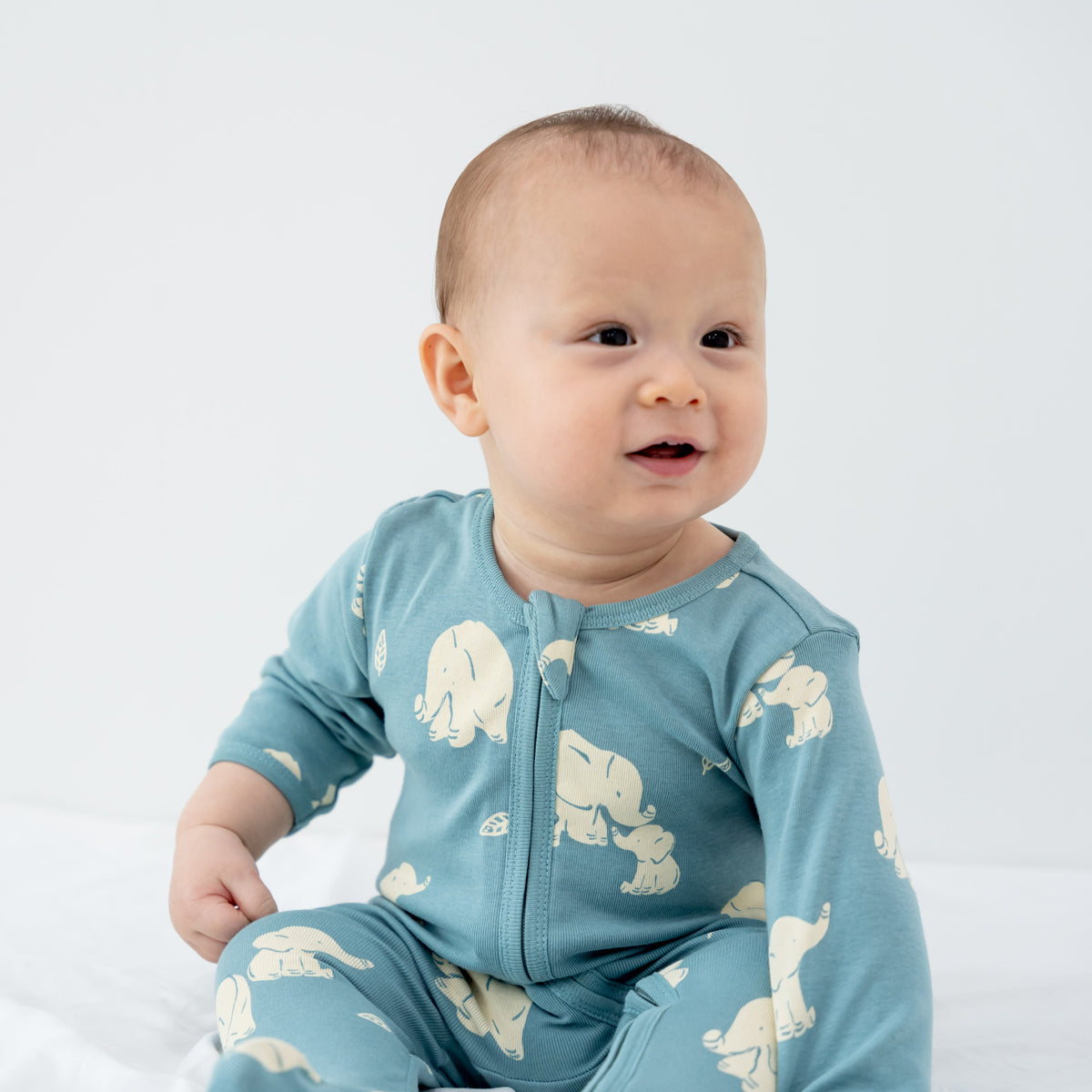 A baby wearing a teal blue zip sleepsuit in elephant print from Le Petit Society.