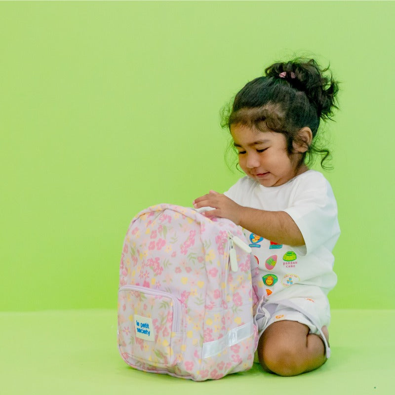 A toddler girl with a purple, floral-print preschool bag.
