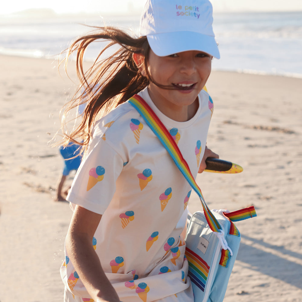 A happy girl carrying a thermal lunch bag with a rainbow print and strap from Le Petit Society.
