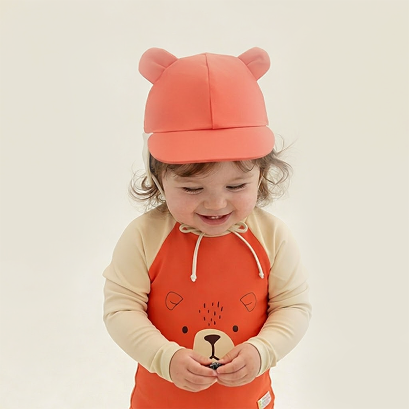 Toddler wearing Le Petit Society Baby Bear Sunhat with matching coral and cream bear rash guard swimsuit with bear face graphic