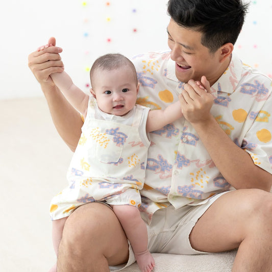 White baby overall from Le Petit Society with colorful dragon playground and sun print in pastel blue, yellow, green, and pink, featuring sleeveless straps and a front pocket.