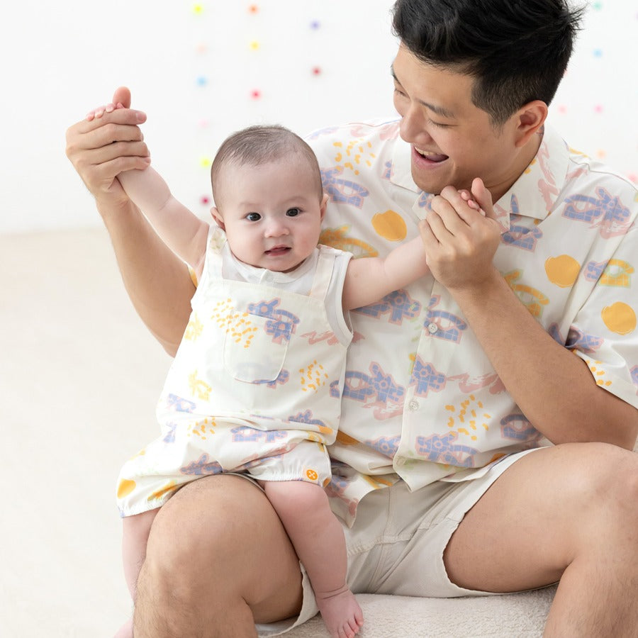 White baby overall from Le Petit Society with colorful dragon playground and sun print, featuring sleeveless straps and a front pocket.