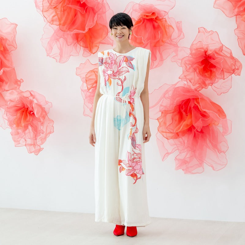 A female model wearing a flowing cream jumpsuit with large pink and blue lotus floral prints from the Le Petit Society x Sheila Sim CNY 2026 collection stands smiling against a white wall decorated with soft coral organza flower accents. She completes the