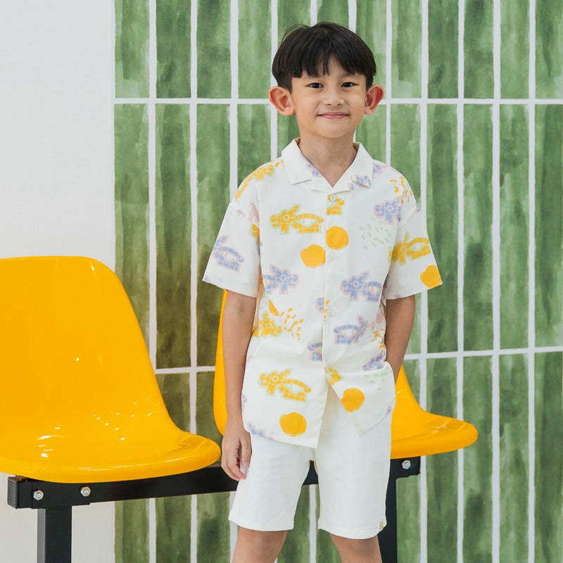 White short-sleeve boys’ button-up shirt from Le Petit Society with camp collar in colorful dragon playground print and sun print in pastel blue, yellow, green, and pink.