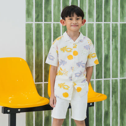 White short-sleeve boys’ button-up shirt from Le Petit Society with camp collar in colorful dragon playground print and sun print in pastel blue, yellow, green, and pink.