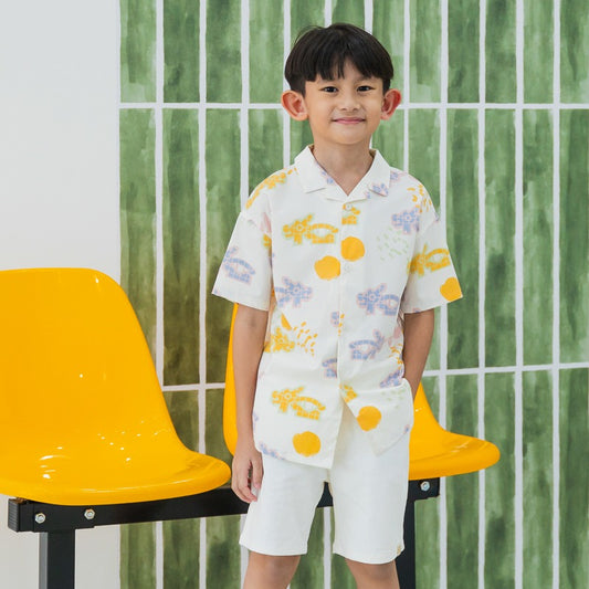 White short-sleeve boys’ button-up shirt from Le Petit Society with camp collar in colorful dragon playground print and sun print in pastel blue, yellow, green, and pink.