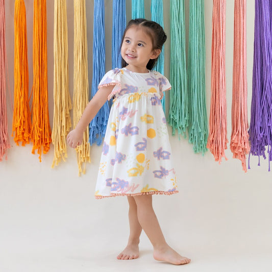 White girl’s flutter-sleeve dress from Le Petit Society with pink pompom trim and colorful dragon playground print and sun print in pastel blue, yellow, green, and pink.