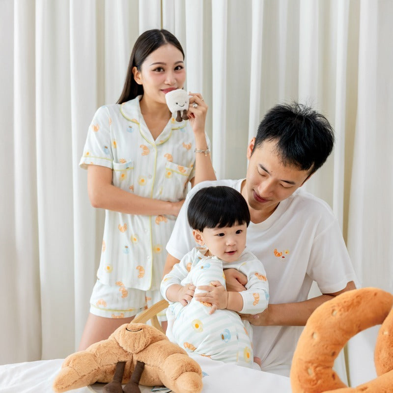 A family of three — mother, father, and baby boy — wearing matching sleepwear from Le Petit Society, featuring a playful print of breakfast food and pastel green stripes.