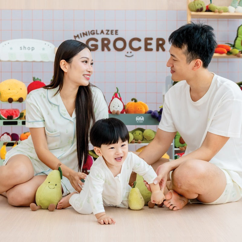 A family of three — mother, father, and baby boy — smiling together while wearing matching outfits from Le Petit Society's sleepwear collection, featuring a print of pastel green pears. 