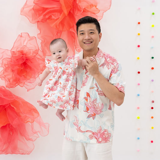 A proud dad holding his little one, both dressed in matching pieces from the Le Petit Society x Sheila Sim CNY 2026 Lotus Collection. The baby wears a flutter-sleeve lotus dress that’s soft, airy and perfect for festive firsts, while dad’s lotus-print shi