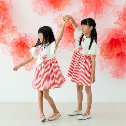 Two young girls wearing matching cream lotus-print tops with red-and-white striped skirt dresses from the Le Petit Society x Sheila Sim CNY 2026 collection hold hands and spin together. They dance in front of a white wall decorated with large coral organz