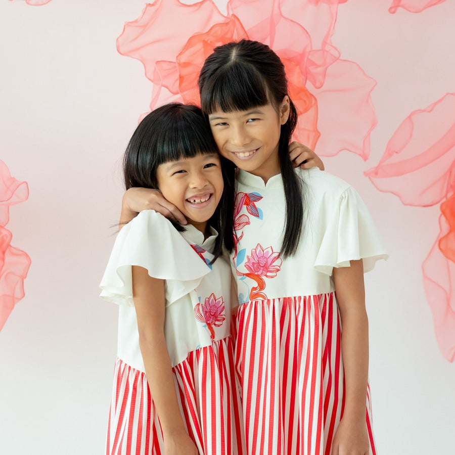 Two young girls stand close together in matching dresses from the Le Petit Society x Sheila Sim CNY 2026 lotus collection. They hug and smile brightly, wearing cream dresses with flutter sleeves, vibrant lotus motifs on the bodice, and bold red vertical-s