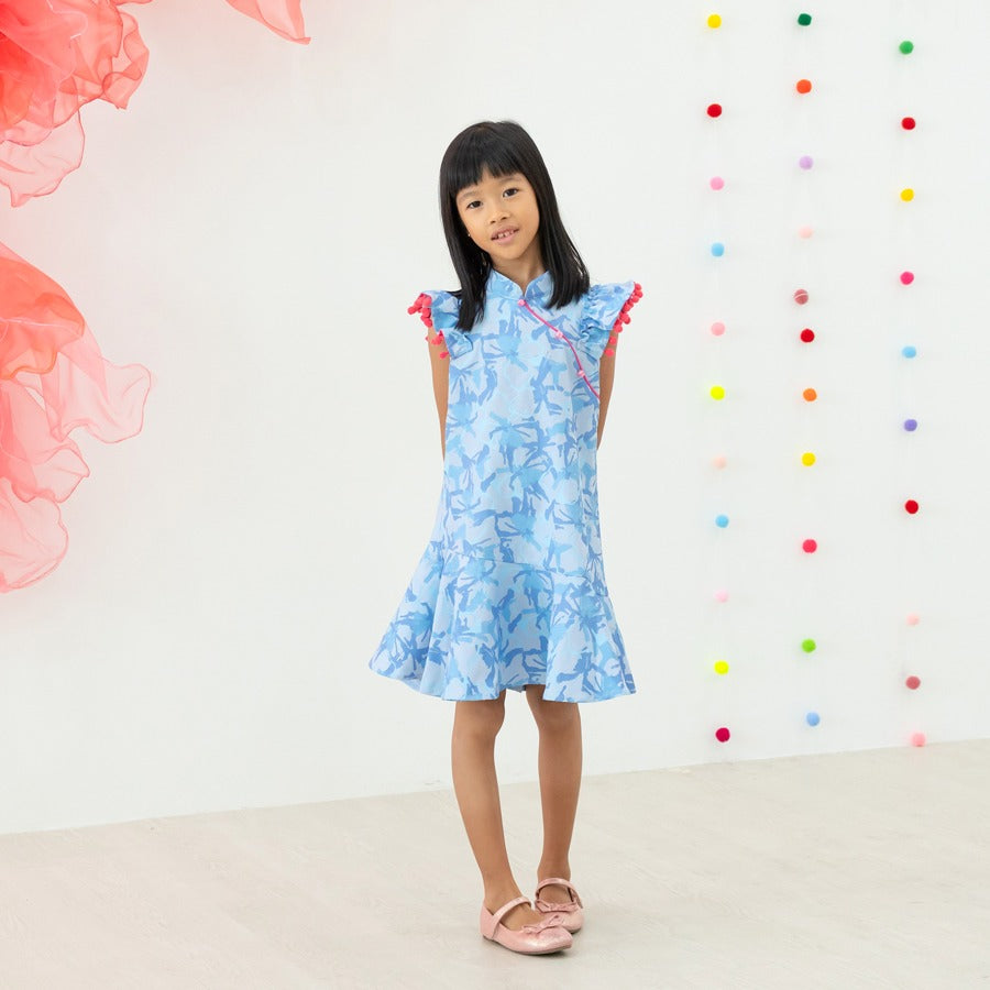 A young girl wearing a light blue ruffle cheongsam dress with pink pom-pom trim from the Le Petit Society x Sheila Sim CNY 2026 collection stands with her hands behind her back. She is smiling softly against a bright backdrop decorated with multicoloured 