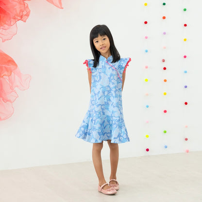 A young girl wearing a light blue ruffle cheongsam dress with pink pom-pom trim from the Le Petit Society x Sheila Sim CNY 2026 collection stands with her hands behind her back. She is smiling softly against a bright backdrop decorated with multicoloured 
